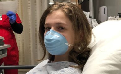A child wearing a face mask and a hospital gown sits upright in a hospital bed and looks at the camera